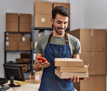 Homem no estoque com alguns produtos em sua mão, sorrindo enquanto olha para o smartphone.