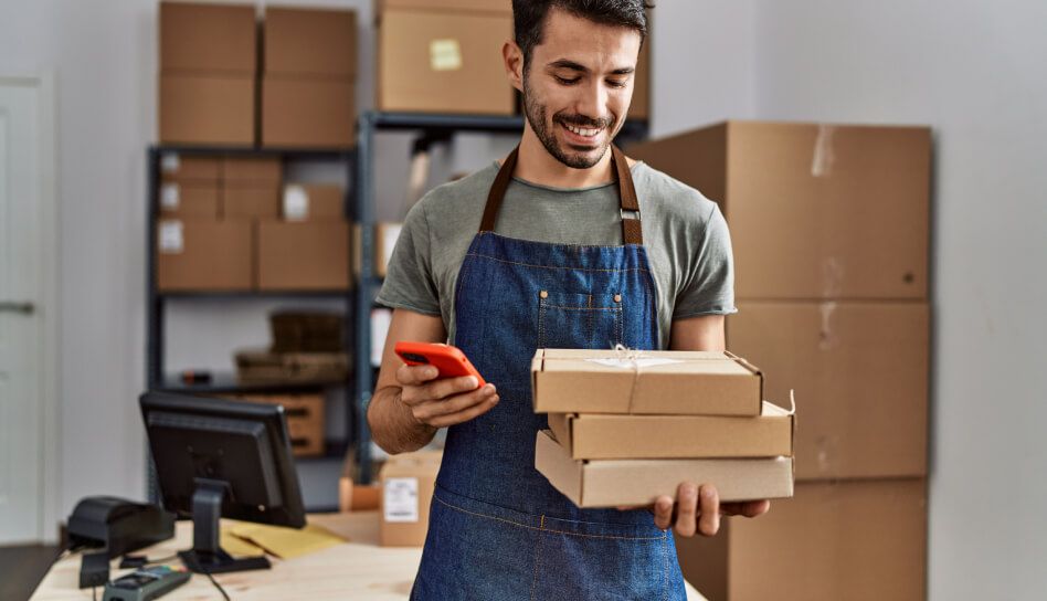 Homem no estoque com alguns produtos em sua mão, sorrindo enquanto olha para o smartphone.